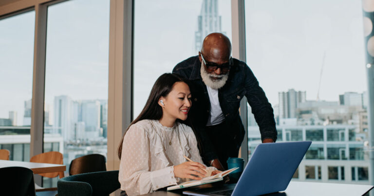 A young woman sits at a desk, taking notes from her laptop. An older man stands behind her, looking over her shoulder at the screen.